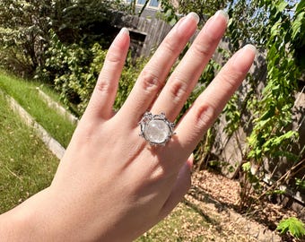 May include: A silver-toned ring featuring a large, round, clear, faceted gemstone at the center. The metal setting has a textured, organic design with small, raised, bead-like accents surrounding the stone, creating a crown-like appearance.