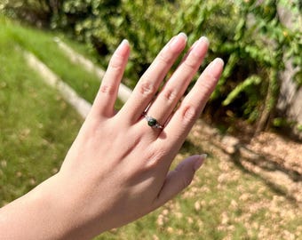 May include: A silver-toned ring featuring a small, round, dark green gemstone set in the center. The band is thin and metallic, with a delicate, textured design on either side of the central stone, creating a simple and refined appearance.
