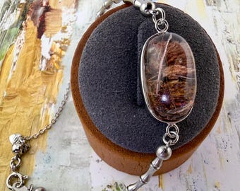 May include: A silver-toned bracelet featuring a central oval-shaped cabochon of clear rutilated quartz with visible brown and golden needle-like inclusions. The chain consists of smooth, segmented metal links resembling bamboo, secured with a lobster clasp.