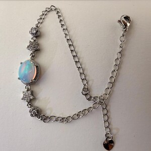 May include: A silver-toned bracelet featuring a central oval-shaped iridescent opal-like stone surrounded by small clear stones. The chain is accented with two star-shaped charms and two round floral-like charms, finished with a lobster clasp and a small heart-shaped charm.