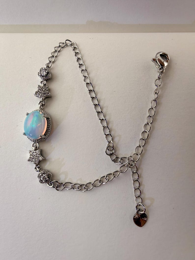 May include: A silver-toned bracelet featuring a central oval-shaped iridescent opal-like stone surrounded by small clear stones. The chain is accented with two star-shaped charms and two round floral-like charms, finished with a lobster clasp and a small heart-shaped charm.