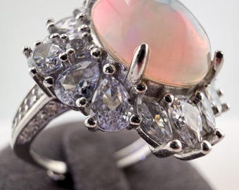May include: A silver-toned ring featuring a large, oval-shaped, iridescent cabochon opal center stone. The opal displays soft pink and white hues. It is surrounded by a cluster of clear, faceted pear-shaped and marquise-cut stones set in silver.