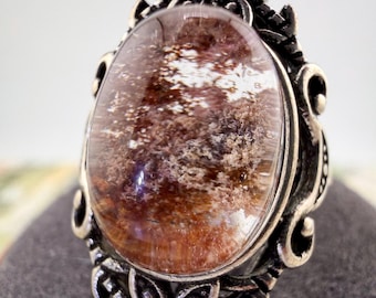 May include: A silver-toned ring featuring an oval-shaped, translucent gemstone with internal reddish-brown and white inclusions. The metal setting is decorated with intricate scrollwork and small flower motifs at the top and bottom edges.
