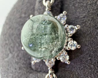 May include: A silver-toned pendant featuring a central, circular, translucent green stone with textured inclusions. The stone is surrounded by a sunburst-style halo of small, clear, round-cut stones, with additional clear stones set along the chain links.