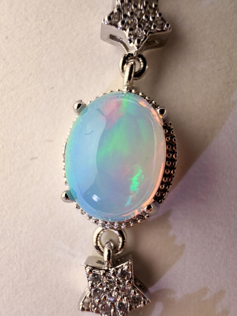 May include: A silver-toned pendant featuring a central oval-shaped iridescent opal cabochon held by four prongs. The setting is accented with two star-shaped charms, one above and one below the opal, both encrusted with small clear stones.