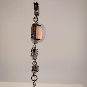 May include: A silver-toned bracelet featuring a central oval-shaped iridescent opal-like stone set in a frame of small clear stones. The chain consists of alternating links with small clear stones and simple circular metal connectors.