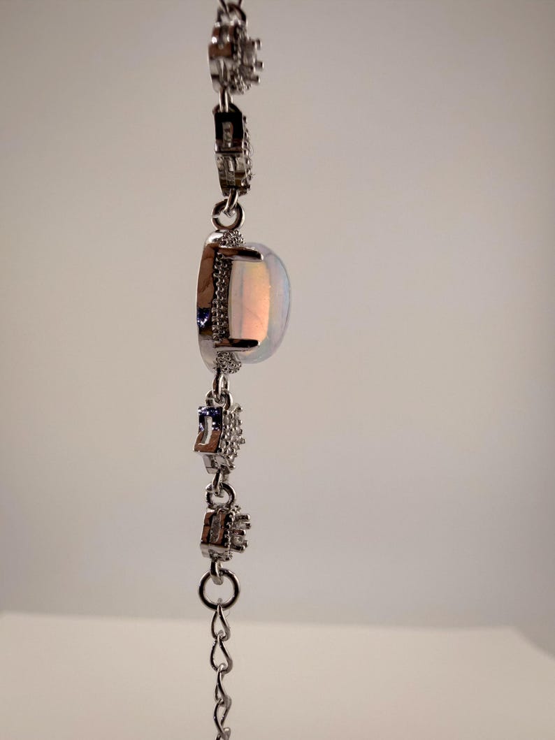May include: A silver-toned bracelet featuring a central oval-shaped iridescent opal-like stone set in a frame of small clear stones. The chain consists of alternating links with small clear stones and simple circular metal connectors.