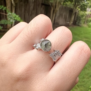 May include: An open-style silver-toned ring featuring a circular, speckled grey stone setting on one end and a cluster of small, clear, faceted stones on the other end. The band is metallic and polished, with additional small clear stones along the sides.