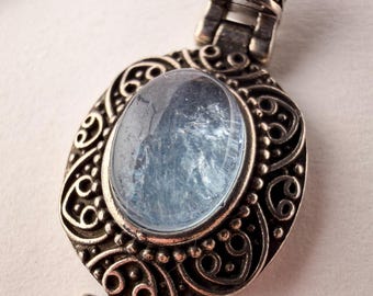 May include: A silver pendant necklace featuring an oval, light blue, translucent stone set in an intricate, oxidized silver frame with scrollwork and beaded details. The pendant is attached to a silver chain with matching decorative metal links.