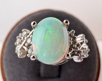 May include: A silver-toned ring featuring a large, oval-shaped cabochon opal with iridescent green and blue flashes. The central opal is held by four prongs and is flanked on both sides by a cluster of small, round, clear faceted accent stones.