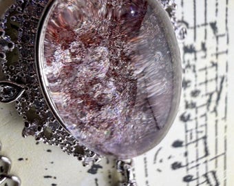 May include: A silver-toned pendant necklace featuring a large, oval-shaped, translucent stone with internal reddish-brown and metallic inclusions. The stone is set in a decorative silver frame adorned with small, clear, sparkling stones.