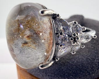 May include: A silver-toned ring featuring a large, oval-shaped, translucent cabochon gemstone with internal golden and grey inclusions. The setting is accented with a cluster of small, clear, marquise and round-cut stones on the side.