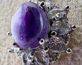 May include: A silver-toned pendant necklace featuring a large, oval-shaped, deep purple cabochon stone at the center. The stone is surrounded by a halo of clear, pear-shaped and round faceted stones set in a metallic frame with a delicate chain.
