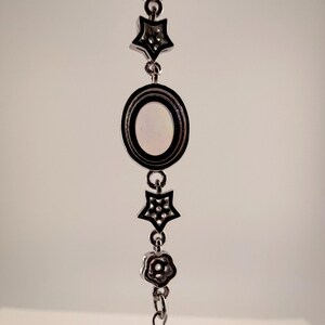 May include: A silver-toned metal bracelet featuring a central oval charm with a smooth, light-colored inlay, flanked by two star-shaped charms with small embedded stones and two flower-shaped charms, all connected by a delicate link chain.