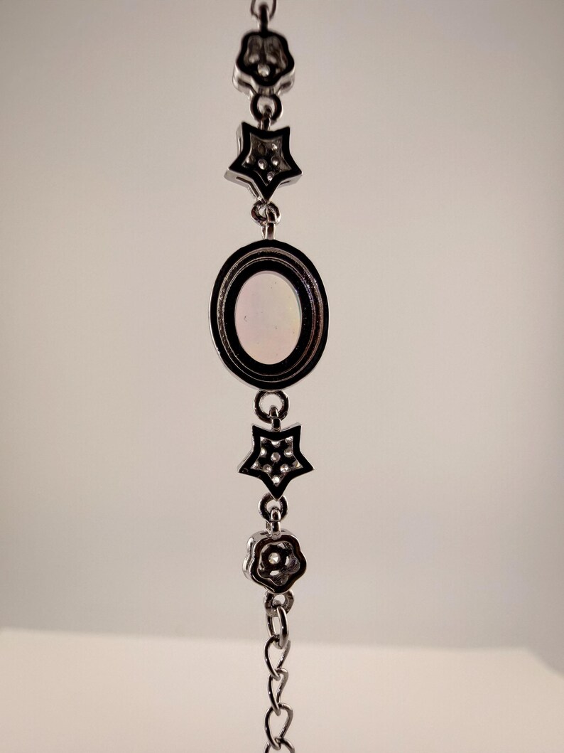 May include: A silver-toned metal bracelet featuring a central oval charm with a smooth, light-colored inlay, flanked by two star-shaped charms with small embedded stones and two flower-shaped charms, all connected by a delicate link chain.