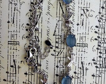 May include: A silver-toned bracelet featuring a series of metallic bow-shaped links alternating with three smooth, oval-shaped, translucent light blue cabochon stones. The bracelet includes a lobster claw clasp and an adjustable chain extension.