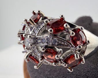 May include: A silver-toned ring featuring a cluster of oval-cut deep red garnet-colored stones arranged in a floral-inspired pattern, accented by small clear white stones set along the central metal bands and prongs holding the red gems.