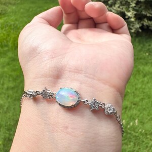 May include: A silver-toned bracelet featuring a central oval-shaped iridescent opal stone surrounded by a halo of small clear stones, accented with star-shaped charms and additional clear stones along the delicate metal chain links.