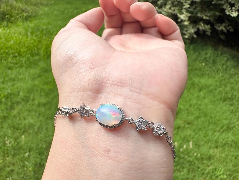 May include: A silver-toned bracelet featuring a central oval-shaped iridescent opal stone surrounded by a halo of small clear stones, accented with star-shaped charms and additional clear stones along the delicate metal chain links.