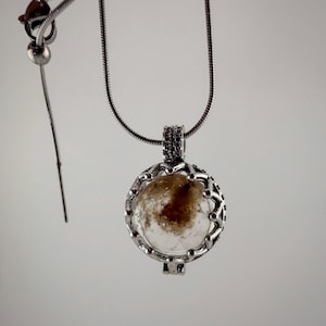 Puede incluir: Un collar con colgante de tono plateado que presenta una piedra redonda y transparente con inclusiones de color marrón oscuro, engastada en un marco de metal decorativo festoneado. El colgante cuelga de una cadena de serpiente fina.