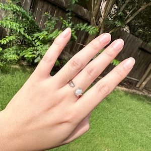 Puede incluir: Un anillo de estilo abierto que presenta una pequeña piedra ovalada iridiscente engastada en una banda metálica. El anillo tiene un espacio entre el engaste de la piedra y un extremo metálico liso y redondeado, creando un diseño abierto.