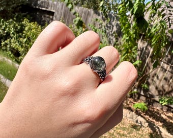 May include: A silver-toned ring featuring a round, translucent green gemstone with dark inclusions. The metal band displays an intricate, textured design with small, raised floral or leaf-like details surrounding the central stone setting.