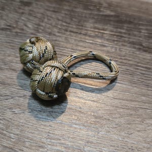 Può includere: Un nodo decorativo, probabilmente un pugno di scimmia, realizzato con corda beige e nera. Il nodo è collegato a un anello della stessa corda. L'oggetto è appoggiato su una superficie di legno.