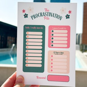 May include: A white paper task list titled The Procrastination Pile with floral accents. It features three sections: a dark green column labeled Sadly, I really must do, a pink box labeled I guess I could do, and a light pink box labeled If I have time.