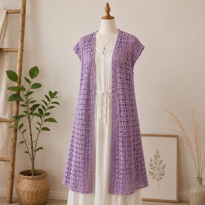 Puede incluir: Un chaleco de ganchillo color lavanda abierto por delante que presenta un delicado patrón circular repetitivo en toda la tela. La prenda no tiene mangas y tiene un dobladillo recto que cae por debajo de la cintura, sobre un vestido blanco.