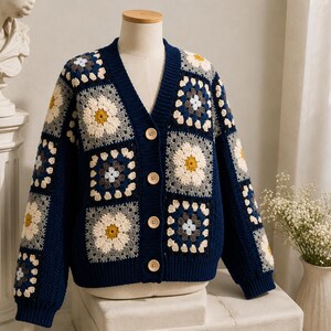 May include: A navy blue V-neck cardigan featuring a crochet granny square pattern with floral motifs in cream, yellow, and grey. The garment has long sleeves, a ribbed hem and cuffs, and is fastened with four light-colored circular buttons.