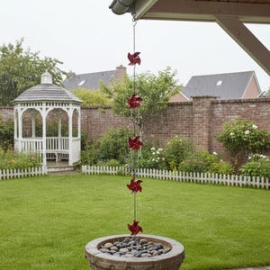 Op de afbeelding: Een rode windmolen regenketting hangt aan een dakgoot, met water dat in een stenen bekken met kiezels stroomt. De ketting hangt boven een groen gazon, met een witte gazebo en een bakstenen muur op de achtergrond.