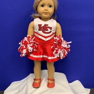 May include: A doll wearing a red and white cheerleader outfit with the letters "KC" on the front. The doll is wearing red glitter shoes and holding red and white pom-poms.