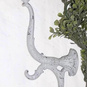 May include: A distressed white metal wall hook with a decorative design. The hook has a curved shape with three points for hanging items. The finish has a worn, vintage look. A green leafy plant is visible in the background.