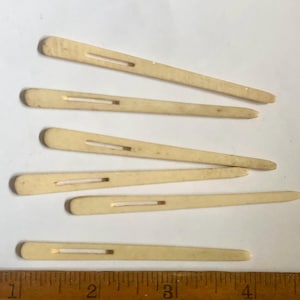 May include: Six vintage ivory-colored hair pins with a rectangular slot in the center. The pins are arranged on a white surface next to a wooden ruler with inch markings. The pins are slightly different lengths.