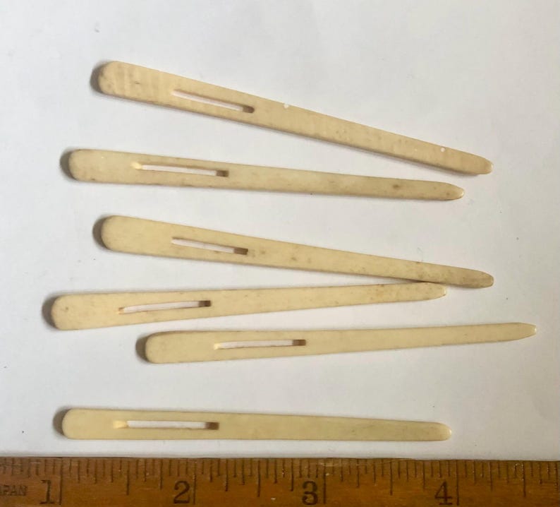 May include: Six vintage ivory-colored hair pins with a rectangular slot in the center. The pins are arranged on a white surface next to a wooden ruler with inch markings. The pins are slightly different lengths.