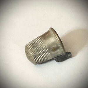 May include: A silver thimble with a textured surface and a small black plastic piece attached to the side.