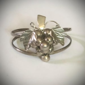 May include: A silver-toned cuff bracelet featuring a central cluster of metallic spheres arranged to resemble a bunch of grapes, accented with two polished metal leaves and a small stem detail attached to a double-banded open cuff design.