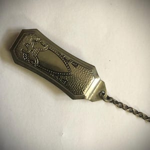 May include: A silver-toned metal decorative piece with an engraved design and a chain. The rectangular piece has a textured surface and a shield-like emblem with a floral and animal motif. The chain is made of small, interlocking links.