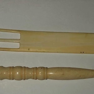 May include: Two antique ivory-colored tools. One is a long, flat tool with a central prong and a curved end. The other is a small, pointed tool with a decorative handle. Both tools appear to be made of bone.