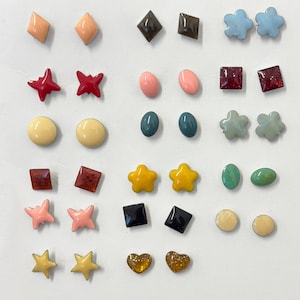 May include: A collection of colorful stud earrings in various shapes, including diamonds, stars, squares, and circles. The earrings come in shades of red, pink, yellow, blue, and green. Some have glitter accents.