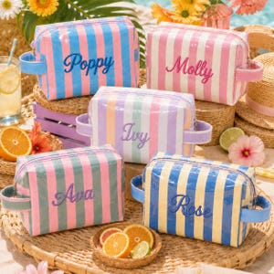 May include: Five colorful, striped cosmetic bags with embroidered names: Poppy, Molly, Ivy, Ava, and Rose. Each bag has a different color scheme, including pink, blue, green, and yellow stripes. The bags have matching handles and are displayed on a woven tray.