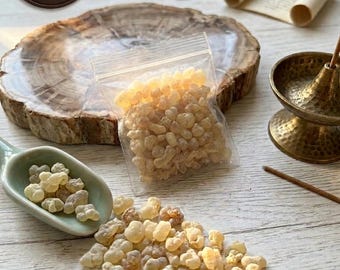 May include: A clear plastic bag filled with pale yellow frankincense resin, accompanied by a light green ceramic spoon containing more resin, a brass incense burner, and a circular brown tag labeled Danat Salalah The Pearl of Oman.