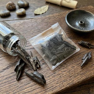 May include: A clear glass jar tipped on its side, spilling dark, jagged agarwood chips onto a wooden surface. A small, transparent plastic bag filled with similar dark wood pieces sits nearby, alongside a dark, circular incense burner.
