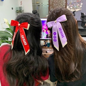 May include: Two hair bows attached to dark hair. One bow is red with white trim and features the text ARIRANG and a circular logo. The second bow is light purple with white trim, also displaying the text ARIRANG and a circular logo.