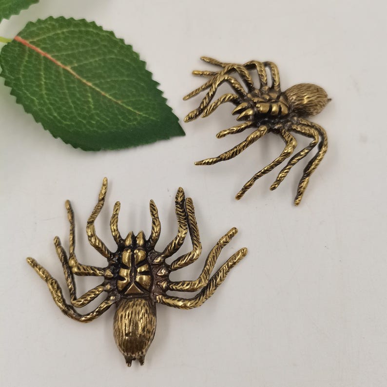 May include: Two metallic brass-toned spider ornaments featuring detailed textured legs and segmented bodies. Each spider has eight legs extending outward from a central thorax and abdomen. The finish is a muted antique gold color with dark highlights.