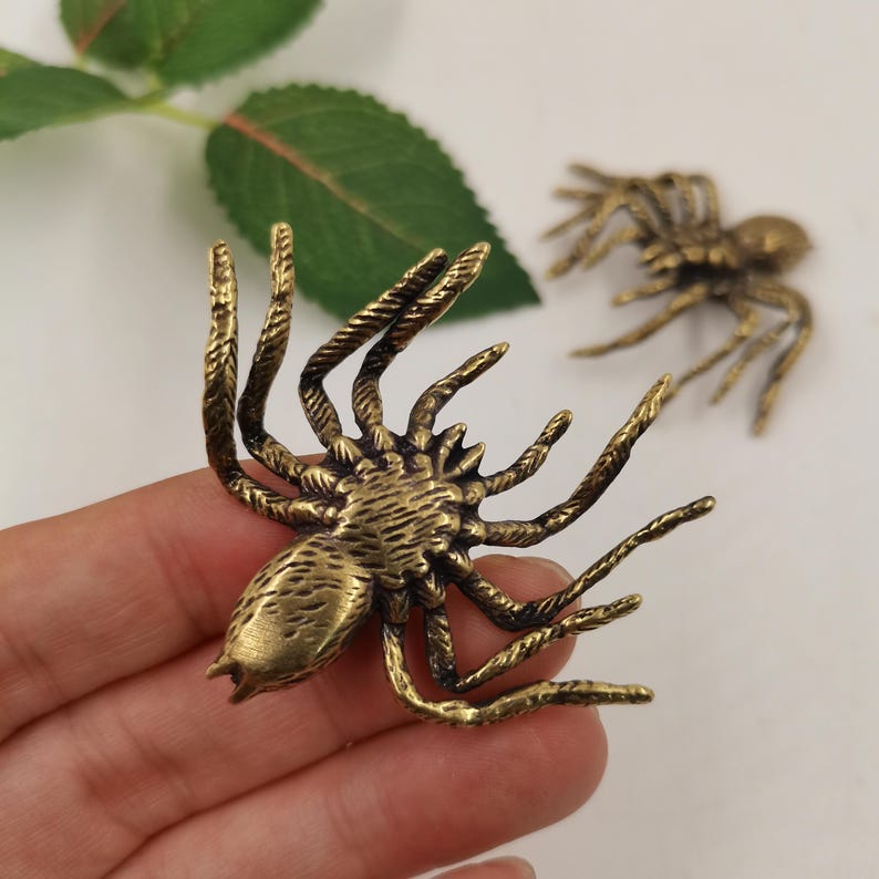 May include: Two brass-colored metal spider ornaments with textured bodies and eight spindly legs. The spiders feature a metallic finish with dark antiqued detailing in the crevices of the legs and abdomen, showcasing a realistic arachnid shape.