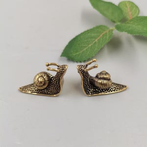 Peut inclure: Une paire de figurines d'escargots en métal couleur laiton avec une surface texturée et pointillée et une coquille en spirale. Chaque escargot est représenté de profil avec des pédoncules oculaires, arborant une finition métallique.
