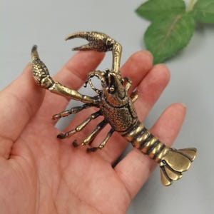 Puede incluir: Una pequeña y detallada figura de latón que representa un cangrejo de río o una langosta. La escultura de metal presenta un caparazón texturizado, pinzas prominentes levantadas, múltiples patas articuladas y una cola segmentada en abanico.