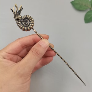 May include: A bronze-toned metal hair stick featuring a detailed ammonite fossil topper with a spiral shell design and tentacle-like protrusions. The long, slender shaft of the hair accessory has a consistent twisted, spiral texture throughout its length.