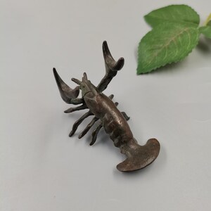 Puede incluir: Una pequeña figura de metal de color bronce oscuro que representa una langosta con las pinzas levantadas, un cuerpo segmentado y una cola ancha. La escultura presenta una superficie texturizada y desgastada con sutiles reflejos verdes.